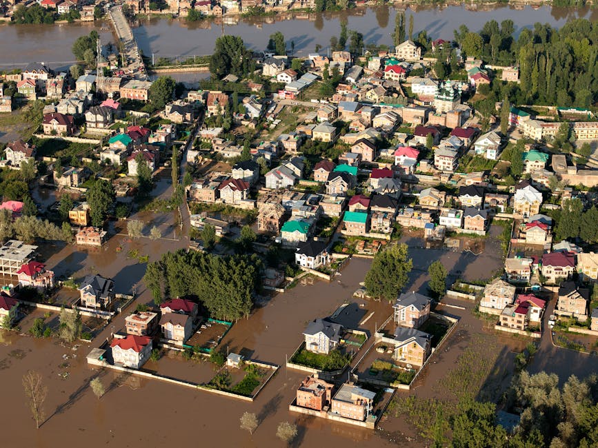 Aerial view of a residential neighborhood with numerous houses surrounded by floodwaters following a recent flood event. The flooded streets and yards are visibly submerged, with water covering large portions of the properties and roads. Some houses have clear garden borders, trees, and driveways, while others are partially flooded, with water reaching up to the lower windows or garden fences. The scene is illuminated by daylight, highlighting the extent of flooding across the area. The image showcases the urgent need for professional surface cleaning and sanitisation, which Putney Carpet Cleaners offers during emergencies like flood aftermaths in Putney. The overview emphasizes the importance of efficient flood response and hygiene restoration for affected homes.