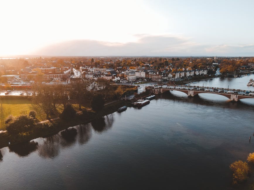 An aerial view of Putney Bridge spanning the River Thames in the late afternoon, with the riverside flats and historic buildings along the shoreline. The scene features a mix of residential and commercial structures, some with pitched roofs and others with flat rooftops, set amidst trees showing autumn foliage. The river reflects the sky above, which is partly cloudy with warm sunlight casting a gentle glow over the scene. A stone bridge with multiple arches supports vehicle traffic, while boats are docked along the riverbank. The overall image showcases a clean, picturesque riverside scene typical of Putney, emphasizing the importance of surface cleaning and maintenance for properties in this area, as highlighted by Putney Carpet Cleaners' expertise in domestic cleaning and surface sanitisation services.