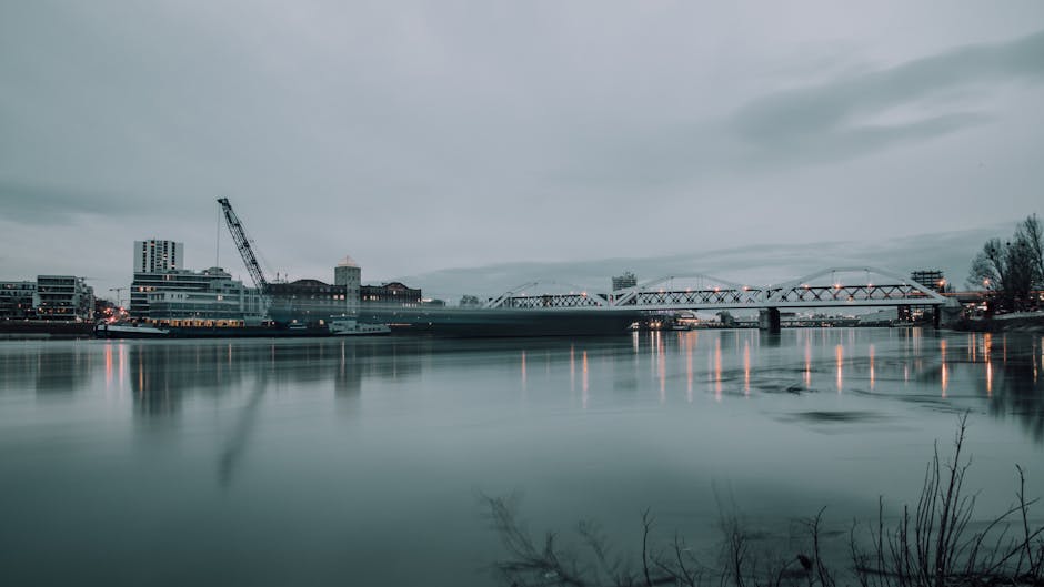 A panoramic view of the riverside area near Putney Bridge showing a calm, reflective river with a smooth surface. In the background, there are modern buildings and a construction crane on the left side, indicating ongoing development. To the right, a bridge spans across the water, with its metal framework illuminated by streetlights, casting warm reflections onto the river. The sky is overcast with gray clouds, creating a muted, neutral lighting atmosphere. The shoreline in the foreground has some bare branches and sparse vegetation. This image exemplifies the cleanliness and maintenance of riverside areas, relevant to surface cleaning and environmental hygiene, as highlighted by Putney Carpet Cleaners in their tips for maintaining riverside flats.