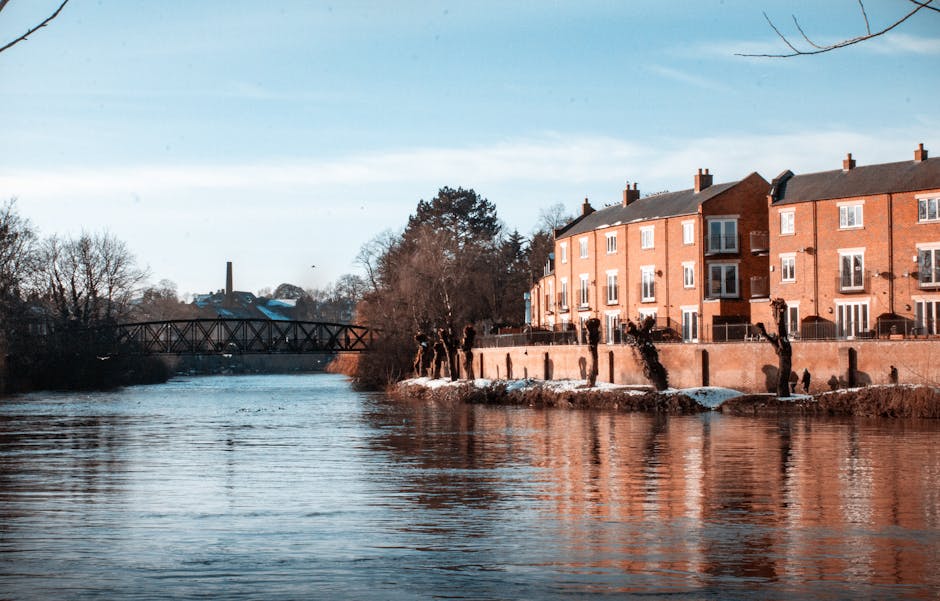 An aerial view of Putney Bridge spanning the River Thames in the late afternoon, with the riverside flats and historic buildings along the shoreline. The scene features a mix of residential and commercial structures, some with pitched roofs and others with flat rooftops, set amidst trees showing autumn foliage. The river reflects the sky above, which is partly cloudy with warm sunlight casting a gentle glow over the scene. A stone bridge with multiple arches supports vehicle traffic, while boats are docked along the riverbank. The overall image showcases a clean, picturesque riverside scene typical of Putney, emphasizing the importance of surface cleaning and maintenance for properties in this area, as highlighted by Putney Carpet Cleaners' expertise in domestic cleaning and surface sanitisation services.