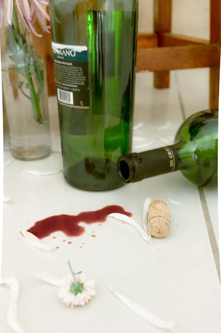 A close-up view of a white countertop in a domestic kitchen or dining area, featuring a spilled red wine stain with a surrounding trail of drips and splashes. The scene includes several empty green glass wine bottles, one of which is partially visible laying on its side, with corks placed nearby. In the background, there are some decorative glass vases and a wooden table, all illuminated by ambient indoor lighting. The overall impression highlights the importance of surface cleaning and stain removal, consistent with the services offered by Putney Carpet Cleaners, as seen on their page for red wine stain removal for Putney homes.