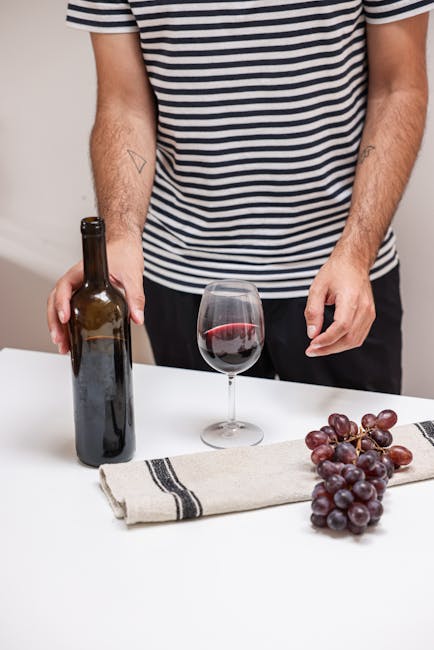 Close-up of a person's torso and arms, wearing a black and white striped T-shirt, standing at a white table. On the table, there is an empty wine bottle, a glass of red wine with a slight spill on the rim, a bunch of purple grapes resting on a beige cloth with black stripes. The background features a plain, light-colored wall. The image conveys a casual domestic setting related to surface cleaning and maintenance, consistent with services offered by Putney Carpet Cleaners for domestic cleaning and stain removal, such as red wine stains.