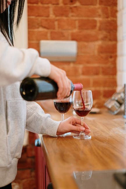 A close-up view of a white countertop in a domestic kitchen or dining area, featuring a spilled red wine stain with a surrounding trail of drips and splashes. The scene includes several empty green glass wine bottles, one of which is partially visible laying on its side, with corks placed nearby. In the background, there are some decorative glass vases and a wooden table, all illuminated by ambient indoor lighting. The overall impression highlights the importance of surface cleaning and stain removal, consistent with the services offered by Putney Carpet Cleaners, as seen on their page for red wine stain removal for Putney homes.
