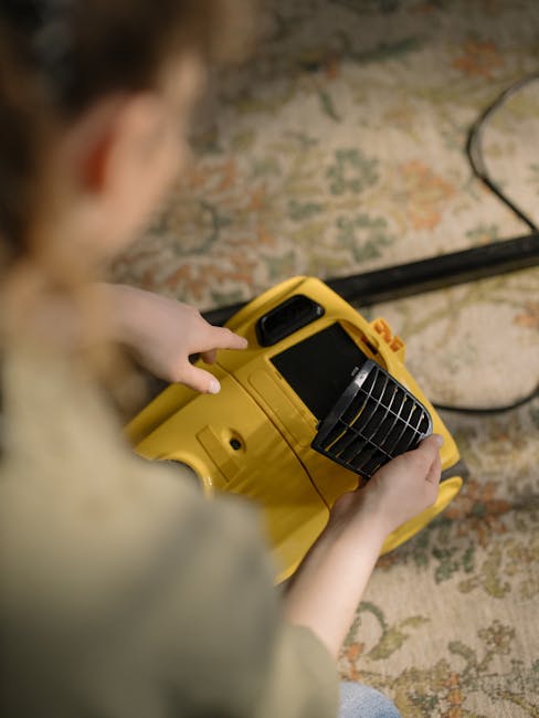 Close-up of a modern vacuum cleaner with a transparent dust container filled with dirt and debris, connected to a flexible, patterned hose, positioned on a pink carpeted floor in a residential room. T