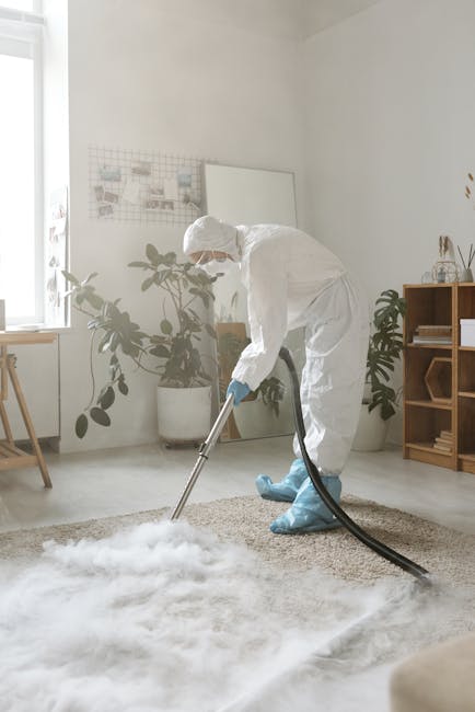 A professional cleaner dressed in a white protective suit, gloves, and a face mask is using a portable vacuum and steam cleaning equipment to deep clean a light beige carpet in a bright, well-lit living room. The room features white walls, a large window allowing natural light, and decorative indoor plants in white pots. A wooden shelving unit with various decor items is visible in the background. The scene emphasizes thorough surface cleaning and sanitisation carried out by Putney Carpet Cleaners, highlighting their expertise in domestic cleaning services to maintain hygiene and cleanliness in residential spaces.