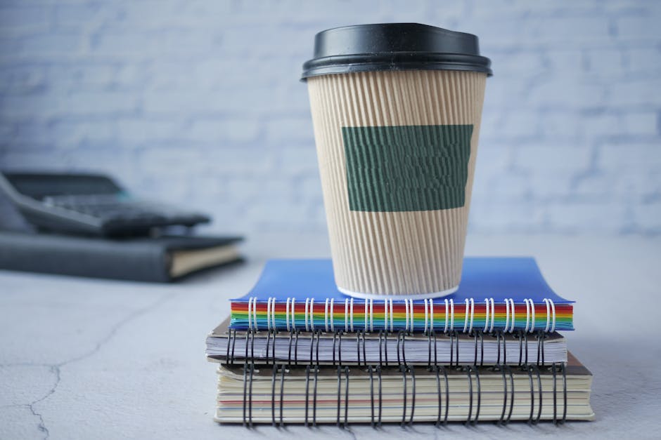 A takeaway coffee cup with a black lid and beige corrugated sleeve is positioned on top of a stack of spiral-bound notebooks and colored files, placed on a white surface. The background features a blurred light blue brick wall, suggesting an office or workspace setting. The scene depicts a tidy and organized environment, suitable for discussions related to cleaning or waste disposal. The presence of cleaning products or tools is not visible, but the neatness aligns with professional cleaning standards explained on PutneyCarpetCleaners.com, particularly concerning surface cleaning and sanitisation linked to the Wandsworth Council carpet disposal rules in Putney.