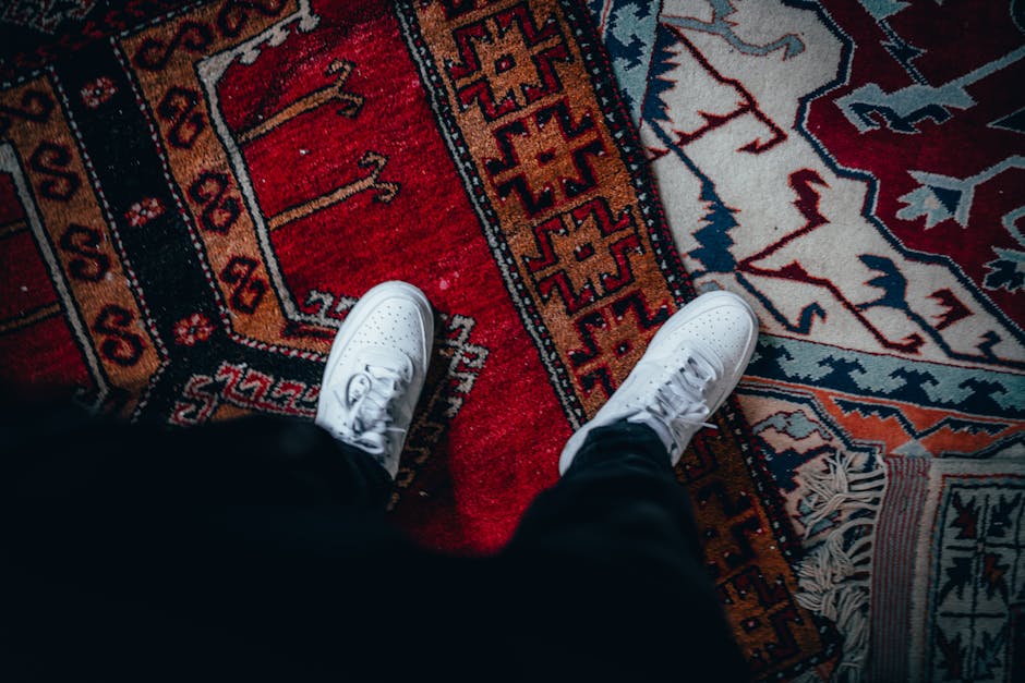 A top-down view of a person standing on a traditional patterned area rug with intricate geometric designs in shades of red, orange, blue, and cream. The individual is wearing white sneakers and dark pants, and the surface of the rug appears clean and well-maintained. The lighting highlights the vibrant colors and detailed motifs of the rug, emphasizing domestic surface cleaning and deep cleaning practices. This setting illustrates a professional cleaning service, such as Putney Carpet Cleaners, focusing on achieving hygienic and visually appealing home environments.